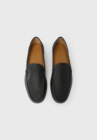 Ζευγάρι μαύρα δερμάτινα loafers χωρίς κορδόνια με καφέ εσωτερικά πέλματα, στοιχισμένα προσεκτικά δίπλα δίπλα πάνω σε λευκό φόντο.