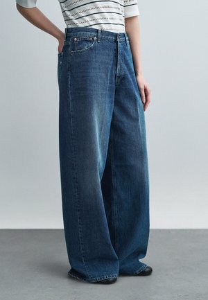 Persoon die wijde donkerblauwe spijkerbroeken en een gestreept cropped top draagt, staand met één hand in de zak.