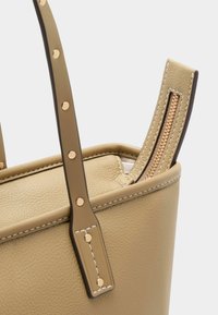 Bézs színű bőr kézitáska sima textúrával, dupla felső fogantyúval, aranyszínű fém alkatrészekkel és díszítő varrással. Oldalán cipzár részlettel.