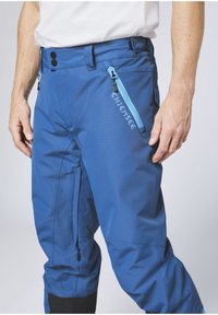 Blue ski pants made of waterproof fabric. Features a visible side pocket with a blue zipper, adjustable waistband, and black ankle cuffs.
