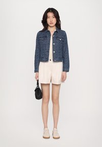 Jeansjacke mit strukturiertem Muster, hellblauer Farbe, silbernen Details, kombiniert mit einem cremefarbenen Oberteil, beigen Shorts und weißen Sneakers.