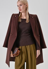 Femme aux cheveux blonds portant un manteau marron foncé sur un pull assorti, un pantalon large olive, des boucles d'oreilles en or en forme de créoles, et tenant une pochette à carreaux marron.