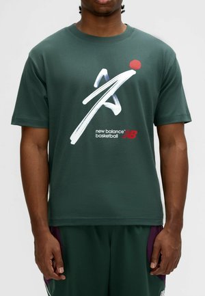 Mand iført en mørkegrøn New Balance basketball-t-shirt med en hvid og rød grafik på forsiden.