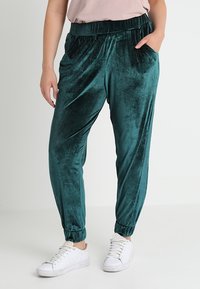 Pantalons de jogging en velours de couleur bleu sarcelle, avec une taille élastique, des poches latérales et des poignets resserrés. Assortis à des baskets blanches pour un contraste.