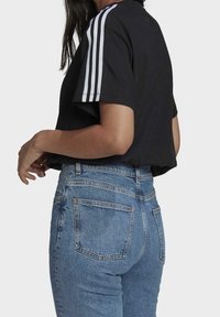 Vrouw in een zwart T-shirt met witte strepen op de mouwen en hoog-waisted blauwe denim jeans, zichtbaar van achteren en van opzij.