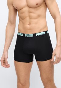 Άνδρας μοντέλο που φοράει μαύρα μποξεράκια Puma με ανοιχτό μπλε λογότυπο Puma στη μέση, στέκεται μπροστά σε απλό φωτεινό φόντο.