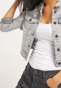 Kvinna som bär en ljusgrå denimjacka över en vit linne och svarta jeans, med ena handen som rör vid sitt hår och den andra i fickan.