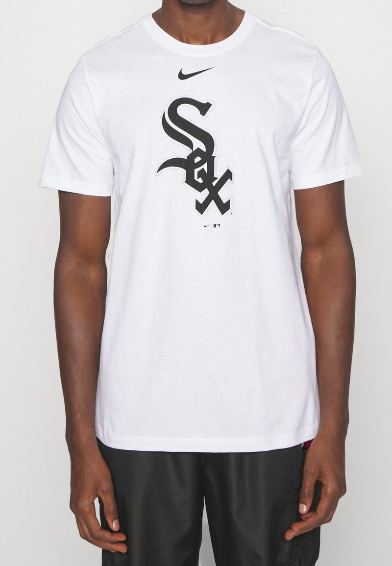 Homme portant un T-shirt Nike blanc avec un logo "Sox" noir centré sur le devant, associé à un pantalon noir, debout devant un fond uni.