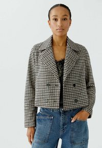 Kürzer geschnittene Hahnentritt-Jacke in Schwarz und Creme mit doppelt geknöpften Knöpfen, Reverskragen und strukturiertem Schnitt, kombiniert mit blauen Jeans.