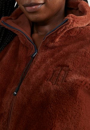 Gros plan d'une personne portant une veste polaire marron zippée avec une capuche et un logo embossé sur la poitrine.