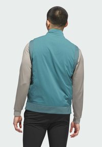 Gilet sans manches en teal avec une texture lisse, doté d'un col haut et d'une taille élastique, superposé sur un haut beige à manches longues.
