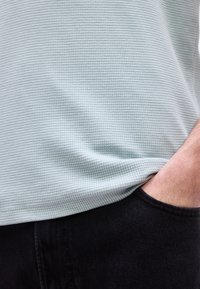 Κοντινό πλάνο ενός ατόμου που φοράει ένα ανοιχτόχρωμο πουκάμισο με υφή, με το δεξί του χέρι τοποθετημένο άνετα στην τσέπη μαύρου παντελονιού.