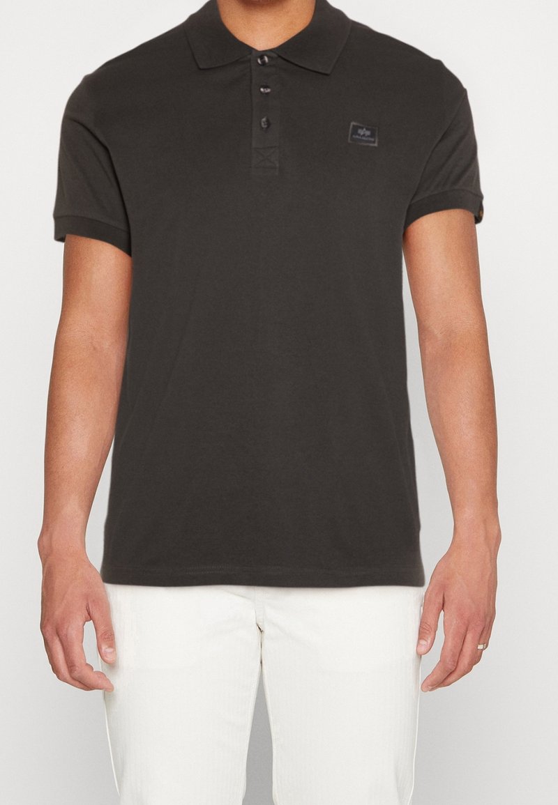 Homme portant un polo noir uni à manches courtes avec trois boutons et un pantalon blanc, debout devant un fond clair.