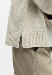 Lys beige strikket genser med ribbet kant og sidespalter, kombinert med lysegrå bukser. Myk tekstur og avslappet design.