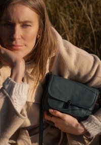 Dunkelgrüne Leder-Umhängetasche mit Klappenverschluss, detaillierten Nähten und verstellbarem Tragegurt, gehalten von einer Person in einem beigefarbenen Pullover.