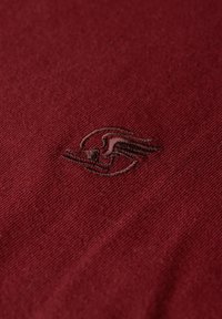 Bordeauxrode stof met een fijne textuur en een klein, donkerbruin geborduurd logo van een gevleugeld figuur, gecentreerd op het materiaal.