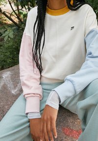 Sudadera a bloques de color con secciones en rosa pastel, azul claro y blanco roto, con un cuello acanalado amarillo y un detalle de logo en el pecho.