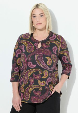 Frau mit blonden Bob-Haaren, die ein schwarzes Oberteil mit rosa und gelben Paisley-Mustern und Dreiviertelärmeln trägt, posiert vor einer weißen Wand.