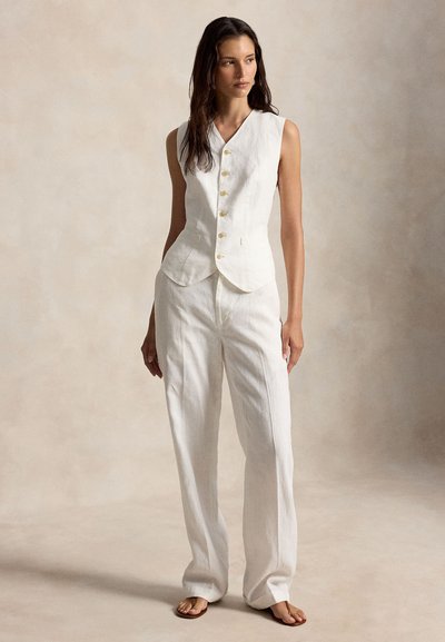 Γυναίκα όρθια, φορά αμάνικο λευκό γιλέκο με κουμπιά, παντελόνι φαρδύ, ταιριαστού χρώματος, και καφέ σανδάλια, μπροστά σε μπεζ φόντο.