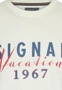 Λευκό μπλουζάκι με στρογγυλή λαιμόκοψη, μπλε σκούρα κεφαλαία γράμματα που σχηματίζουν τη λέξη "SIGNAL", κόκκινα καλλιγραφικά γράμματα "Vacation" και μπλε σκούρα "1967" από κάτω.