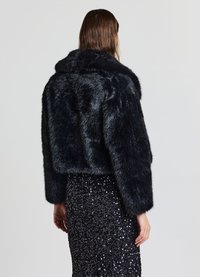 Schwarze, verkürzte Jacke aus Kunstpelz mit weitem Kragen und langen Ärmeln, die eine weiche Textur aufweist. Wird über einem schwarzen, mit Pailletten besetzten Rock getragen.