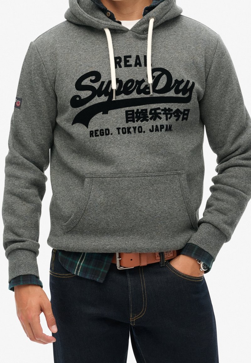 Sudadera con capucha de forro polar gris con estampado gráfico negro, cordones y bolsillo frontal. Llevada sobre una camisa de cuadros verde, pantalones vaqueros oscuros y un cinturón marrón.