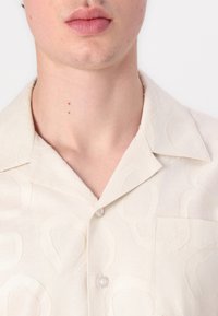 Beige Kurzarmshirt mit strukturiertem Muster, versehen mit einem weichen Kragen, einer Brusttasche und Knopfverschluss. Leichter Stoff mit dezentem Glanz.