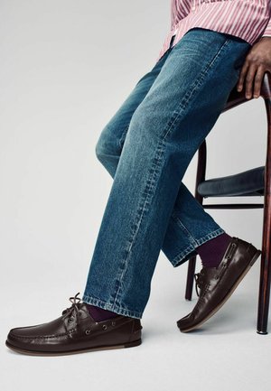 Brune lædersko med snørebånd, metaløjer og struktureret overflade. Båret med blå jeans og mørkelilla sokker, siddende på en træstol.