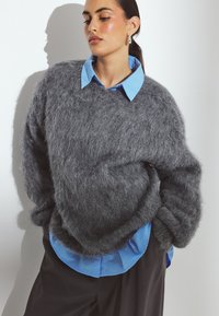 Suéter gris y esponjoso hecho de material suave, con un corte holgado. Debajo, se puede ver parcialmente una camisa de color azul claro con cuello.