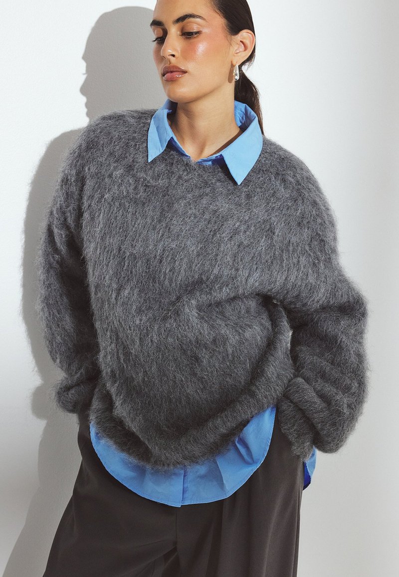 Suéter gris y esponjoso hecho de material suave, con un corte holgado. Debajo, se puede ver parcialmente una camisa de color azul claro con cuello.