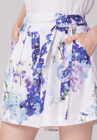 Jupe blanche avec un motif floral dans des tons de bleu et de violet. Elle dispose d'une ceinture à nouer et de poches, avec des détails plissés à la taille.