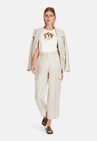 Helle beige Leinenjacke über einem weißen T-Shirt mit floralem Grafikdesign, kombiniert mit weiten beigefarbenen Hosen und braunen Sandalen.