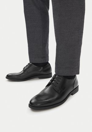 Czarne skórzane eleganckie buty o gładkim wykończeniu, zaokrąglonych noskach i wiązane na sznurowadła, zestawione z ciemnoszarymi teksturowanymi spodniami.