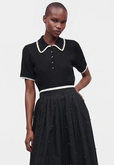 Femme portant un haut noir à manches courtes avec bordure blanche et boutons, associé à une jupe noire avec de petits détails en perles et une ceinture blanche.