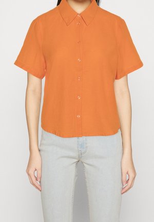 Frau trägt ein kurzärmliges oranges Hemd mit Knöpfen, gesteckt in hellblaue Jeans, steht vor einem einfachen weißen Hintergrund.