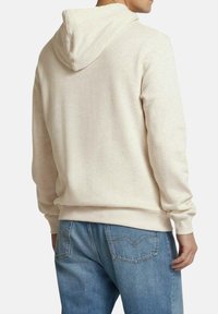 Sudadera de color beige con capucha ajustable, textura suave y puños de canalé. Combinada con jeans de mezclilla de color azul claro, mostrando detalles en la parte trasera.