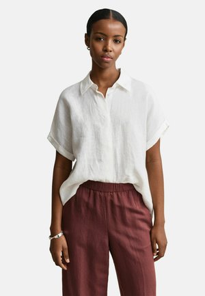Vrouw die een loszittend wit overhemd met korte mouwen draagt, gestoken in hooggetailleerde bordeauxrode broek, staand tegen een effen achtergrond.
