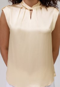 Satin-beige bluse med høy krage, med en nøkkelhull hals og myke folder ved skuldrene. Glatt tekstur, casual passform.