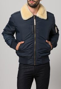 Veste bomber bleu marine avec un col en fausse fourrure beige, fermeture éclair, poches latérales et poignets et ourlet côtelés. Tissu en nylon lisse.