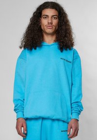 Sudadera azul claro con un bolsillo frontal, con un logotipo impreso en negro en el pecho. Fabricada con una tela suave y texturizada con capucha ajustable.