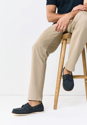 Mann trägt beige Hose und dunkelblaue Loafers, sitzt auf einem Holzschemel vor weißem Hintergrund, Hände auf dem Knie ruhend.