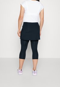 Kvinna klädd i en vit kortärmad topp, marinblå kjol, svarta korta leggings och vita sneakers med rosa och lila accenter, stående med ryggen mot oss.