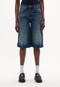 Denim kuloti ar tumši zilu krāsu, vidēja augstuma jostasvieta, priekšējām kabatām un saplēstu malu, apvienoti ar melniem loafers zābakiem un gaišām zeķēm.