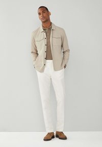 Mann steht mit Händen in den Taschen, trägt eine beigen Hemdjacke, ein taupefarbenes Poloshirt, weiße Hosen und braune Wildlederschuhe vor einem schlichten grauen Hintergrund.