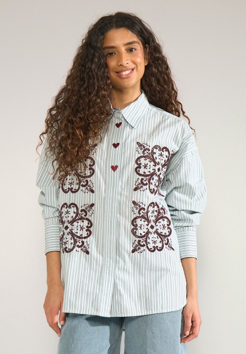 Jeune femme aux longs cheveux bouclés porte une chemise rayée avec une broderie florale rouge et des boutons en forme de cœur assortis, associée à un jean bleu clair.