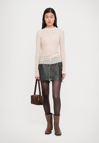 Donna con un top lungo con maniche in pizzo color crema, una mini gonna in pelle nera con cerniera frontale, collant scuri, stivali marroni e che tiene in mano una borsa in camoscio marrone.
