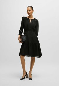 Schwarzes, geschichtetes Kleid mit langen Ärmeln, strukturiertem Stoff und Schlüsselloch-Ausschnitt. Kombiniert mit schwarzen High Heels und einer kleinen Handtasche.