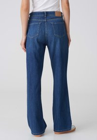 Jean en denim bleu évasé avec taille haute, featuring deux poches arrière et un patch en cuir marron sur la ceinture. Texture douce et lisse.