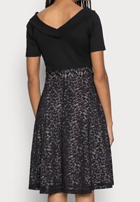 Černé šaty s krátkými rukávy, výstřihem do V na zádech a přiléhavým pasem. Sukně má květinový krajkový překryv s rozšířeným tvarem.