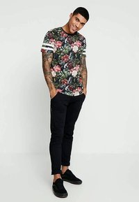 Blomsterdesign t-skjorte med mørk bakgrunn, livlige farger og hvite striper på ermene. Sammen med svarte bukser og joggesko.